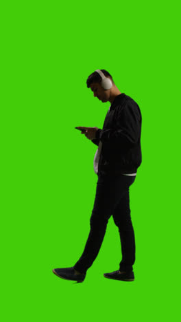 Vertical-Video-Full-Length-Shot-Of-Man-Wearing-Wireless-Headphones-Streaming-Music-From-Mobile-Phone-Against-Green-Screen-2